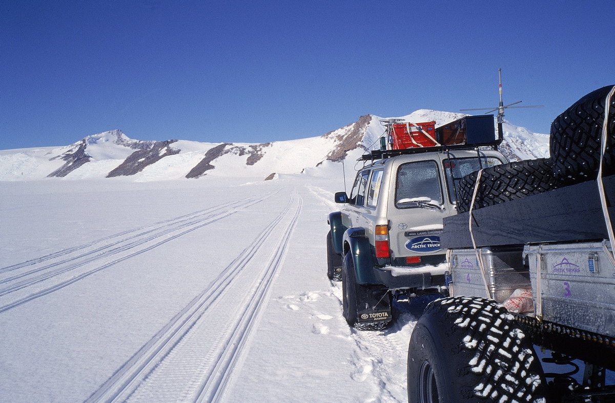 Land Cruiser med sl&auml;p f&ouml;ljer sp&aring;ren &ouml;ver inlandsisen mot bergen i Dronning Maud Land.